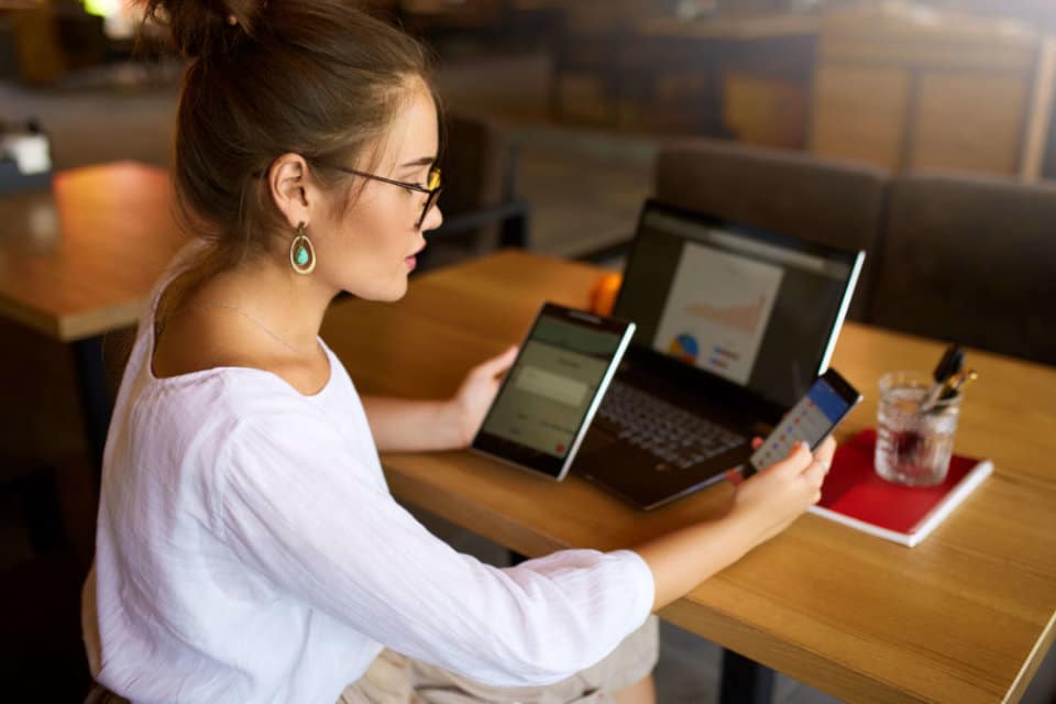 Mixed race woman in glasses working with multiple electronic internet devices. Freelancer businesswoman has tablet and smartphone in hands and laptop on table with charts on screen. Multitasking and telecommuting theme.