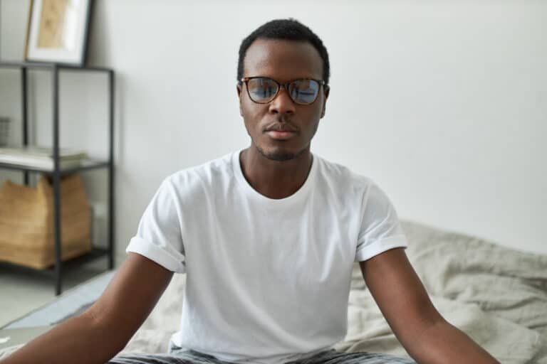 A black gentleman wearing glasses and a white t-shirt sits in meditation with his eyes closed.