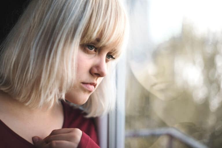 Woman with blonde hair looks out a window in thought.