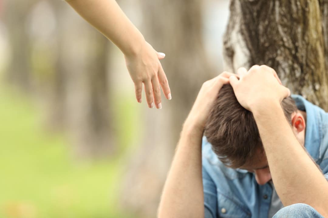 Man leaning against the tree with his head in his hands and a helping hand reaching out to him.