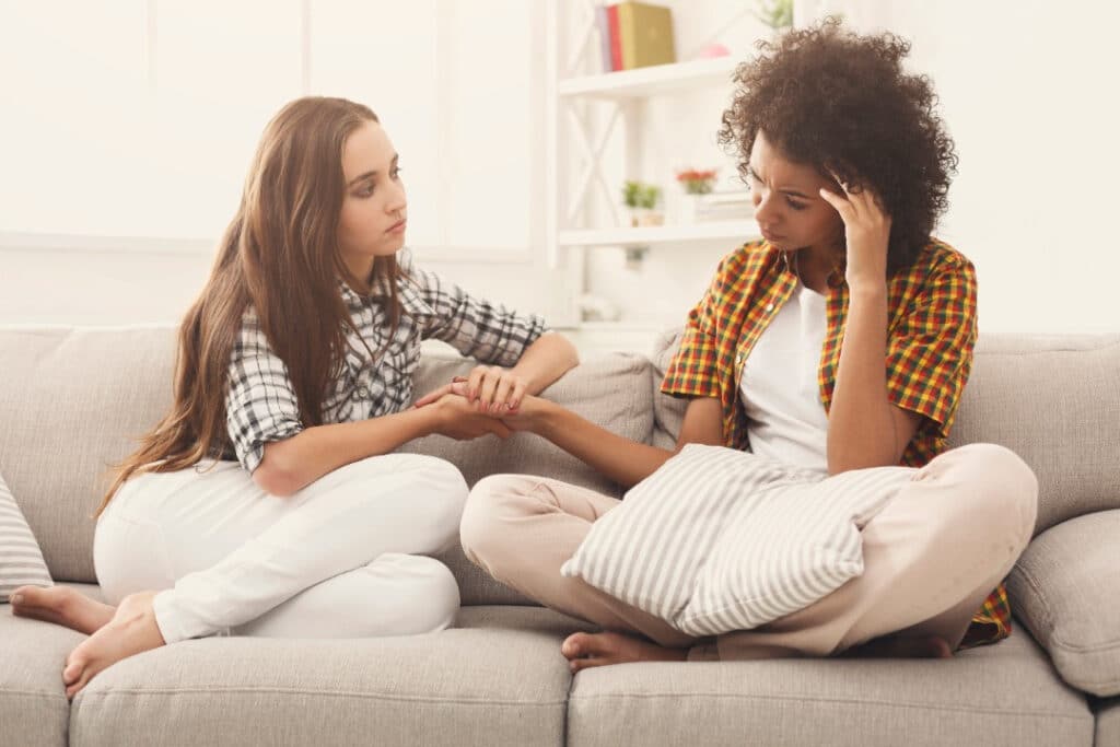 Two friends sitting on the couch. One woman holds the other woman's hand as they talk.